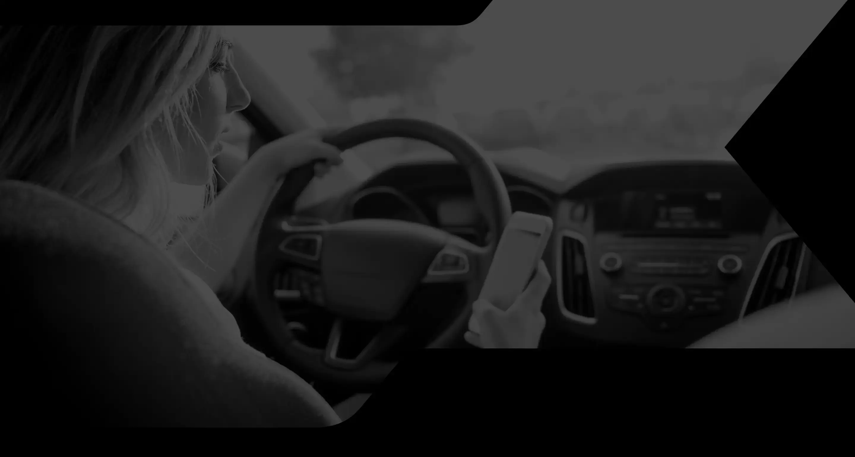 A woman in a car holds a smartphone while driving, with sunlight streaming in. Her focus is on the phone, suggesting distraction at the wheel.