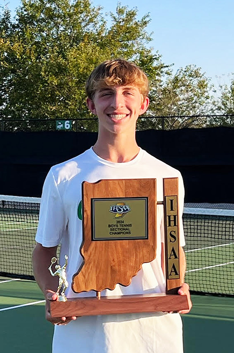 Mitchell, smiling and holding a wooden tennis trophy shaped like Indiana, labeled “2024 Boys Tennis Sectional Champions,” on a sunny outdoor tennis court.
