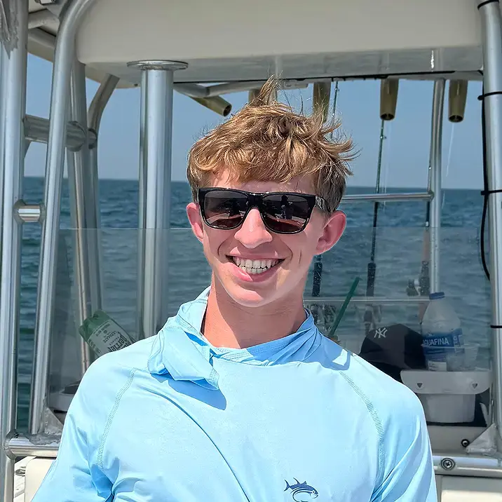 Mitchell in sunglasses and a light blue shirt stands on a boat under a sunny sky, with the water in the background, conveying a joyful mood.